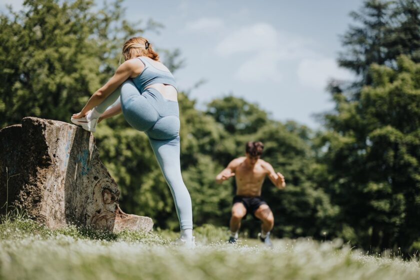 Immagine a tema sesso e sport: due persone fanno esercizi di riscaldamento e calisthenics in un parco, con luce naturale e atmosfera wellness.