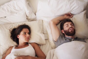 Immagine a tema sesso in coppia: coppia seduta sul letto in pigiama, atmosfera serale stanca ma intima, luce calda e discreta.